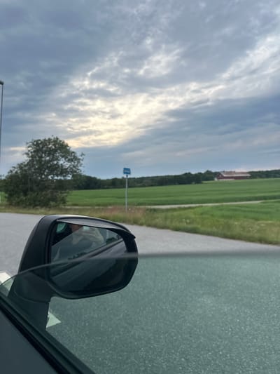 The image shows a side-view mirror of a car with a person's reflection, indicating the photographer is inside the vehicle. Outside the car, a rural landscape unfolds under a cloudy sky. A green field stretches towards a distant red barn. A single tree stands on the left side of the road, and a blue sign is visible in the distance. The road surface appears to be asphalt. The scene is likely located on the outskirts of Slitu, Norway, given the pastoral setting and typical Scandinavian architecture of the barn.