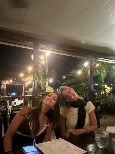 Two women sit at a table in an outdoor dining area. The woman on the left is wearing a black and white lace-trimmed top and a silver necklace. The woman on the right is wearing a white t-shirt and a black scarf. The table is covered with a white tablecloth, and there is a menu, a cell phone, and a glass of water on the table. They are smiling at the camera. String lights and tropical greenery surround them.  The scene is located at coordinates 7.432, -80.189 in Las Escobas del Venado, Panama.