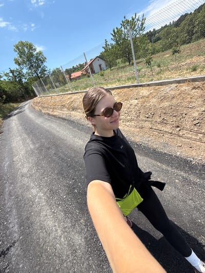 A rotated outdoor selfie captures a person standing on an asphalt path under a bright, clear blue sky with scattered white clouds. The subject, appearing to be female, has light brown hair and wears brown-framed sunglasses, a black short-sleeved top with a visible "KO/" logo, black leggings, and a neon green fanny pack or bag worn around the waist. White socks and athletic shoes are visible on one foot.
To the subject's left, a chain-link fence separates the paved path from a grassy area with some dry patches and several trees. In the background, beyond the trees, a building with a distinctive red-tiled roof can be seen, suggesting a rural or semi-rural setting within Grocka Urban Municipality, Serbia. The sun is high, casting strong shadows, indicating it is bright daylight, likely midday or early afternoon. The overall scene suggests a pleasant day for outdoor activity.