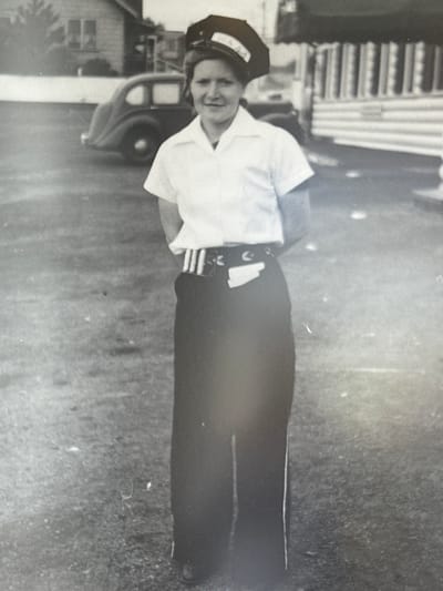 A black and white photograph depicts a woman standing outdoors in what appears to be the Township of Langley, Canada. She is wearing a uniform consisting of a white short-sleeved shirt, a dark skirt or pants, and a hat with light-colored lettering. She smiles at the camera. To her left is a car, and behind her, a building. The ground appears to be grass or dirt.