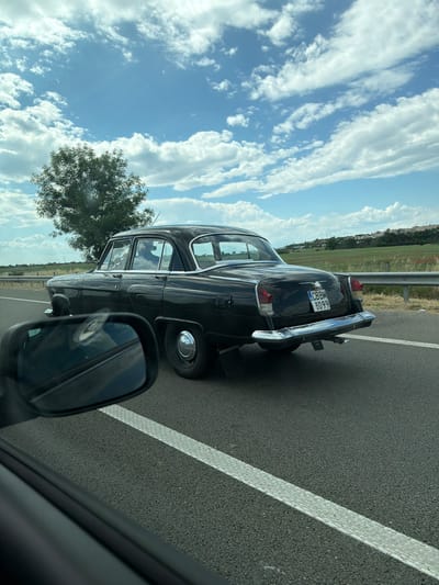 The image shows a black vintage car on a road in what is likely Shishmantsi, Bulgaria, based on the license plate CBBH 8099. The road has a white lane marker. The sky is visible and is mostly blue with white clouds. In the foreground, a car mirror and a portion of a car window are visible. There is a tree and some fields in the background.
