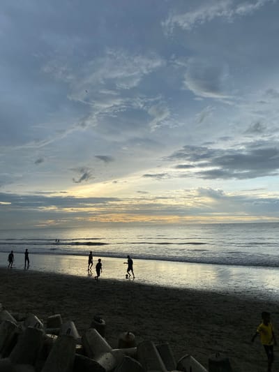 The image captures a beach scene in Bateetimoh, Indonesia, during what appears to be either dawn or dusk, given the soft, diffused light. The sky is filled with a mix of gray and white clouds, with a band of warm, golden light visible on the horizon where the sun is likely low. Gentle waves are rolling onto the sandy shore.
In the mid-ground, several figures are silhouetted against the brighter background, engaged in playing with a ball on the wet sand near the water's edge. These figures appear to be children or young people. Further out in the water, a few other indistinct figures can be seen, possibly swimming or wading.
In the foreground, a collection of large, grey concrete barriers, likely tetrapods or similar coastal defense structures, are arranged on the sand. To the far right of the foreground, a young child in a bright yellow shirt is walking on the sand. The overall atmosphere is peaceful and suggests recreational activity on the beach.