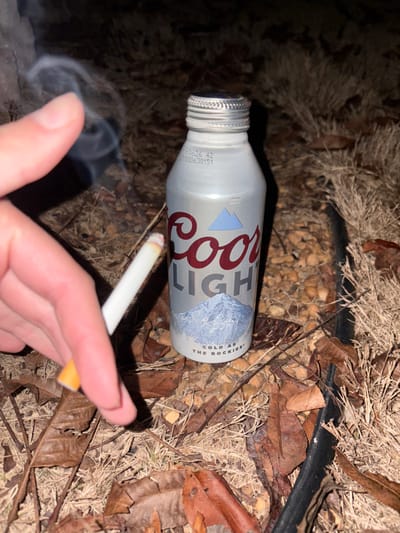 A Coors Light aluminum bottle lies on a bed of dried leaves and small gravel. Next to it, a hand holds a lit cigarette, smoke curling upwards. The backdrop consists of dark, dry vegetation and a thick black cable. The lighting casts shadows, highlighting the details of the discarded bottle and the burning cigarette.