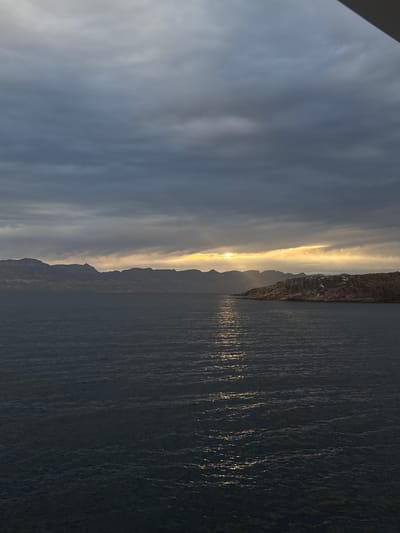 The image captures a landscape at either sunrise or sunset. A large body of water fills the right side of the frame. The water's surface is textured with ripples, reflecting the light from the sun, appearing as streaks. On the left, the sky is filled with clouds, transitioning from darker shades at the top to a brighter, golden hue near the horizon. A mountain range is silhouetted against the bright sky. The location is Unknown, Mexico.