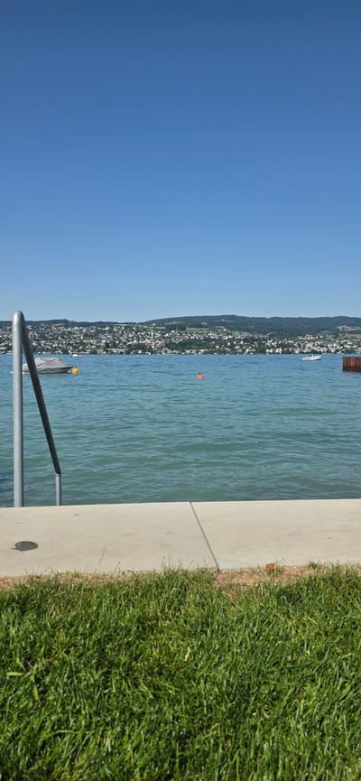 A vertical, eye-level, outdoor medium shot captures a serene scene at the edge of a bright blue lake, likely on a clear, sunny day. In the foreground, lush green grass covers the bottom of the frame, transitioning to a paved concrete walkway. A metal railing with a curved handle is positioned on the left side of the walkway, leading towards the water.
The lake itself is a vibrant turquoise, with gentle ripples on its surface. A small, grey-covered boat is moored in the middle ground on the left, with a single orange buoy floating nearby. Further out, a small orange buoy is visible in the center of the frame. Across the lake, the shoreline is densely populated with buildings, forming a town nestled against rolling, tree-covered hills. The distant horizon is a blend of the town, hills, and the clear blue sky above. This scene is consistent with the shoreline of Lake Zurich in Oberrieden, Switzerland, given the landscape and architectural style. There are no people visible in the frame.