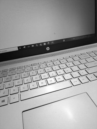 A close-up, monochrome shot focuses on the keyboard and part of the screen of an HP laptop. The QWERTY layout is visible, including letters like Q, W, E, R, and Z, as well as the alt and Windows keys. To the left, the laptop screen displays the Windows taskbar with application icons and the HP logo. The touchpad occupies the upper right corner of the frame. The image highlights the texture of the keyboard and screen, creating a contrast between the tactile keys and the smooth display.