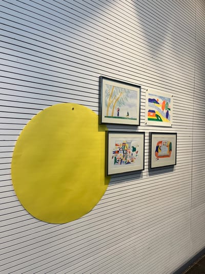 The scene depicts an art display on a horizontally striped white wall inside the Médiathèque de Sélestat, France. A large, bright yellow paper circle, slightly creased, is affixed to the left portion of the wall with a single visible fastener near its top. To the right of the yellow circle, and partially overlapping its edge, are four framed illustrations arranged in a 2x2 grid. All frames are dark and slender. The top-left illustration features two stylized figures under yellow-leafed trees against a light blue sky. The top-right illustration presents a colorful landscape with a red house, rolling hills, and stylized trees. The bottom-left illustration is an abstract composition of various brightly colored shapes and forms. The bottom-right illustration features a stylized figure within a curvilinear, multi-colored interior setting.