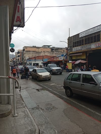 The image depicts a street scene in the city of Los Teques, Venezuela, under an overcast sky. A sidewalk lines the left side of the street, with several pedestrians walking. One individual is pushing a shopping cart, while others are walking in pairs or alone. On the right side of the street, vehicles are parked and driving. A silver hatchback car is prominently featured in the foreground, with a gold sedan in the middle ground. A silver pickup truck is also visible further down the street. Buildings with shops and signs line both sides of the street, suggesting a commercial area. One sign above a building reads "AEROBICS KARATE KOBUDO DE VENEZUELA SPORT KARATE DEFENSA PERSONAL KOBUDO TAEKWONDO CLASES PERSONALIZADAS KUNG FU Y MEDITACION BAIOTERAPIA". Another sign displays "MIRANDINOS". The street itself appears wet, possibly from recent rain. Electrical wires are strung across the scene above the buildings and street.