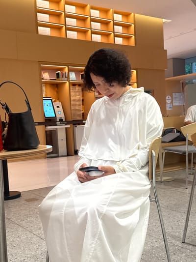 A woman is sitting in a chair and looking at her phone in a room with shelves filled with books. The woman is wearing a white hooded jacket and white pants. The room is lit by warm lighting. The shelves are made of wood and are filled with books. There is a black handbag on a table next to the woman. The scene appears to be in a public space, possibly a library or a cafe, and is located in Xuhui District, China.