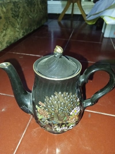 A black glazed ceramic teapot is positioned centrally on a reddish-brown tiled floor. The teapot features a polychrome design on its side, depicting a peacock with an fanned tail and various floral elements in hues of green, gold, brown, pink, and white. It has a curved spout to the left and a C-shaped handle to the right. The lid is black with a circular, gold-toned finial. Minor dust accumulation is visible on the lid and parts of the teapot's surface. A small chip or crack is present at the base of the spout where it meets the main body of the teapot. The reddish-brown floor tiles have visible white grout lines. In the blurred background, a dark green and beige patterned sofa is partially visible on the left, and a light-colored wooden furniture leg and other indistinct objects are visible on the right. This scene is located in Longmiang, Laos.