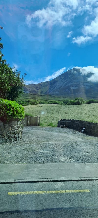 The image depicts a rural landscape in Murrisk, Ireland. The scene is dominated by a steep, grassy mountain slope on the left, partially obscured by dramatic white clouds that hug its peak. The sky is a vibrant blue, suggesting fair weather, though the clouds hint at potential atmospheric changes.

In the foreground, a gravel road runs diagonally across the frame, leading towards the mountain. On the right side of the road, a concrete barrier with a yellow painted line is visible. A low stone wall borders the road and the mountain's base, with a wooden gate set into it. Lush green trees and bushes add texture to the landscape, particularly in the lower-left corner. The fields are a uniform green, suggesting a well-maintained agricultural area.

The perspective implies the photograph was taken from a slightly elevated position, possibly from a vehicle or a viewpoint overlooking the road. The lighting suggests it is daytime, likely late morning or early afternoon, given the brightness and the shadows. There are no people visible, and the focus is entirely on the natural and man-made elements of the rural setting. The overall mood is peaceful and serene, showcasing the rugged beauty of the Irish countryside. No text is visible in the image.