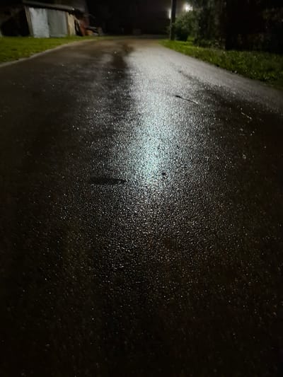 The image depicts a wet asphalt road at night, likely in a rural or suburban area. The road surface is dark and glistening with numerous small water droplets, indicating recent rain. A patch of bright, reflected light, possibly from a streetlamp or vehicle headlights, illuminates a section of the road in the center of the frame. To the left, a grassy verge borders the road, and behind it, a dimly lit structure resembling a shed or outbuilding is visible. The background is dark and out of focus, with hints of vegetation and possibly a pole or post. The overall atmosphere is somber and damp. The location is stated as Novlyanka, Russia. There are no people or overt human activity visible in the image.