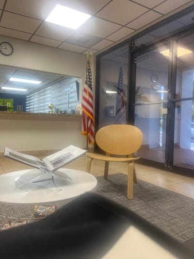 The image depicts an interior scene, possibly a public building like a library or office, in Miami Shores, United States. The perspective is from a low angle, looking upwards and sideways, as the image is rotated 90 degrees counter-clockwise.
In the foreground, a round white table is partially visible, with a display stand holding what appear to be framed documents or photos. The stand is made of clear acrylic. To the left of the table, a patterned rug covers a tiled floor.
Behind the table, a light wood chair with a rounded seat and back is positioned near a large glass-doored entrance. The glass doors reflect the interior and exterior, showing sunlight and trees outside. An American flag is mounted on a pole to the left, partially visible through the glass.
Further inside, a reception desk or counter is visible, with various items on display, including trophies and framed pictures. A clock with Roman numerals is mounted on a cream-colored tiled wall. The ceiling is made of acoustic tiles with inset lighting fixtures. Fluorescent lights are also visible on the ceiling.
The scene appears to be during the daytime, with natural light streaming in from the entrance. There are no people visible in the image. The overall impression is one of a clean, organized, and professional setting. Visible text includes the numbers on the clock and possibly text on the displayed documents.