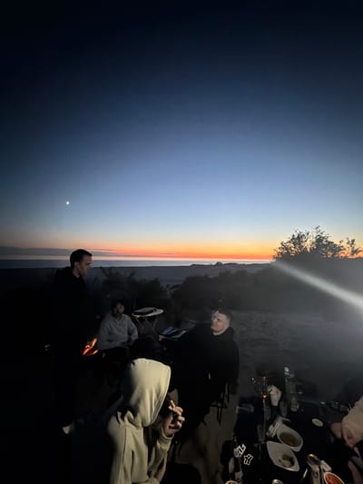 At dusk, a group of people are gathered outdoors, possibly in Baltiysk, Russia, near a fire. The sky transitions from deep blue to shades of orange and pink near the horizon, with a sliver of the moon visible. The foreground is dark, with a campfire providing a warm glow. Several individuals are visible, some silhouetted against the fire and the twilight sky. One person in the foreground is wearing a light-colored hooded sweatshirt and appears to be holding something. To the right, a table is set with food and drinks, suggesting a meal or gathering. The background shows low-lying vegetation and a distant landscape.