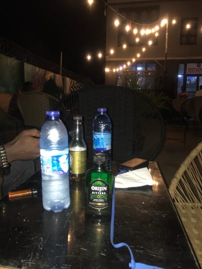 Three bottles line a shiny black surface alongside an orange lighter. Two are filled with clear liquid, while one is a green-labeled "Orijin Bitters" bottle. A dark wicker chair sits beside the table. A hand and beaded bracelet are visible. Overhead, string lights illuminate a building facade with circular motifs and a "Restaurant" sign. A white napkin and blue strap are also on the table.