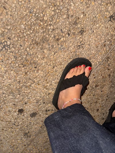 A human left foot, adorned with red nail polish on the toes, is depicted wearing a black platform sandal with a ruffled fabric strap. A multi-strand ankle bracelet with pink, white, and metallic beads is visible on the ankle. The foot and lower portion of a dark blue denim pant leg rest on a paved surface consisting of small, light brown and grey pebbles embedded in concrete. The background shows more of this pebbled ground. The specific location within Azor, Israel, cannot be determined from the visual information provided.