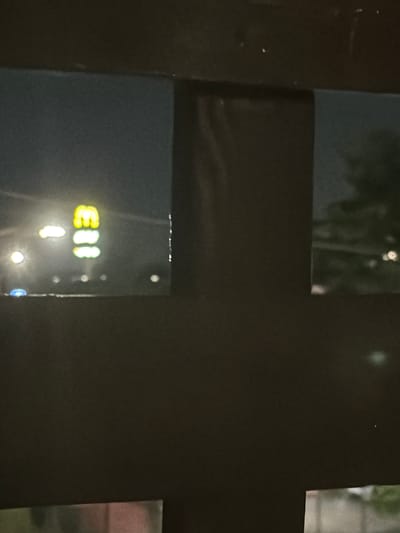 An out-of-focus, low-light image captured at night shows a dimly lit scene through what appears to be a fence or barrier. In the left portion of the frame, through an opening, a brightly lit, vertical sign with three illuminated yellow shapes is visible. These shapes resemble the letter "M" stacked vertically, with the top one slightly wider and angled, and the bottom two more rectangular. To the left of this sign, a blurred white light is present, and further to the left, a faint blue light can be seen. Below these lights, a row of smaller, indistinct lights are scattered horizontally. The background beyond these lights appears dark and hazy, with faint suggestions of trees or foliage to the right. The foreground is dominated by dark, solid structures, likely part of the barrier through which the scene is viewed, creating a segmented perspective. There are no people or clear interactions visible. The image's quality suggests it was taken with a mobile phone camera in a low-light environment. Based on the signage, the location is likely near a McDonald's restaurant in Spinea, Italy.