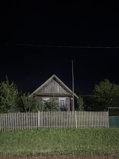An outdoor, eye-level, medium shot captures a wooden house at night, partially obscured by trees and a wooden fence. The house, with a gabled roof and visible windows, is lit from an unseen source, casting shadows. A tall pole with an antenna stands beside the house, and trees flank the structure. In the foreground, a wooden fence runs across the frame, with a patch of grass behind it. The background is a dark, unlit sky. The scene is set in a rural area, likely within the Бучацінскі сельскі Савет of Belarus.