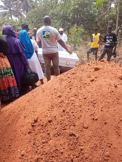 The scene depicts an outdoor burial ceremony in Okpatu-Ukehe, Nigeria. A prominent, irregular mound of reddish-brown, sandy soil occupies the entire foreground, signifying a freshly dug grave. In the mid-ground, a white coffin with gold-colored handles is partially lowered into a rectangular grave opening. Approximately ten individuals are gathered around the grave. A man wearing a light grey t-shirt with a 'STELLAMACTUS' logo on the back and khaki trousers stands directly behind the coffin, facing away from the camera, with his right arm extended towards the coffin, appearing to assist with or direct its placement. To the left of the grave, a woman in a purple head covering and dress holds a black handbag, standing beside another woman in a white dress. To the right of the coffin, two young men observe the process; one wears a yellow 'LAKERS 23' jersey and holds a mobile phone, while the other wears a black 'PERA PAYS 25' t-shirt. The background consists of sparse, dry bushland vegetation, including visible palm trees on the far left, illuminated by bright daylight.