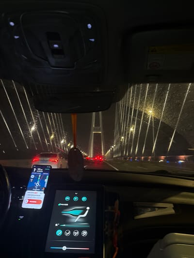The image depicts the interior of a car driving on a bridge at night, with a view of the road ahead through the windshield. The road appears wet, suggesting it might be raining or has recently rained, and there are multiple lanes of traffic visible. Streetlights and vehicle lights create a blurry, streaky effect due to the motion and the wetness on the glass.
In the foreground, on the dashboard, are two electronic devices. One is a large touchscreen displaying what appears to be a car control interface, with icons for AC and other functions. Below it, a smartphone is mounted, showing a navigation app with a map and route information, along with some Chinese text. The phone screen is brightly lit. The car's interior has some illuminated buttons visible.
The overall scene is dark, characteristic of nighttime driving. The lighting is artificial, coming from streetlights and vehicle headlights. The reflection of the lights on the wet windshield adds a dynamic and somewhat abstract quality to the scene. There is a sun visor visible at the top left of the image, with some warning labels, one of which features a symbol of a person with a diagonal line through it, likely indicating a safety warning. There is also a rear-view mirror visible on the left. The location is identified as Xuhui District, China.