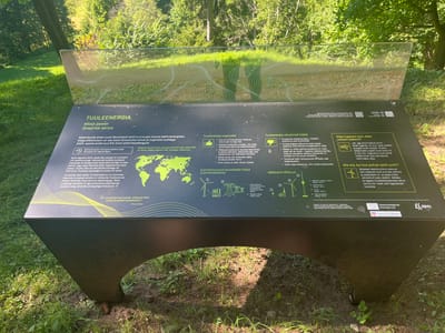 The image shows an outdoor informational display about wind energy in Toila, Estonia. The display is a dark-colored, curved structure with a transparent, reflective top panel that shows a stylized pattern resembling wind currents. The main body of the display features text in Estonian and English, accompanied by graphics.
The text discusses wind power, its benefits, and how electricity is generated from wind. There are illustrations of wind turbines, power lines, and a map of the world highlighting wind energy usage. One section compares wind energy with fossil fuels, listing advantages and disadvantages. Another section explains the process of electricity generation from wind. There are also visual comparisons of different wind turbine sizes and types. QR codes and social media handles ("Nature_of_Estonia", RMK) are visible at the bottom right.
The display is situated outdoors, with lush green grass and trees visible in the background, suggesting a park or natural area. Sunlight filters through the trees, creating shadows on the ground. A fallen pinecone is visible near the base of the display on the ground. The overall setting appears to be a serene, natural environment.