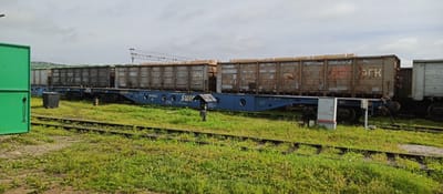 The image depicts a side view of several freight train cars parked on tracks in a grassy, industrial area. The train cars are predominantly a dull, weathered metal color, with some featuring graffiti and faded lettering. The foremost train car is a flatbed with a blue chassis, carrying several large, rectangular cargo containers stacked on top. The containers are also metal and appear to be covered in tan sheeting. The background shows a cloudy sky and a distant industrial structure, possibly a loading crane or gantry. A bright green structure, also industrial in nature, is partially visible on the far left of the image. The ground is covered in green grass and patches of dirt. The scene appears to be a railyard or industrial loading facility. The location is identified as Pogranichny, Russia. There are no people visible in the image.