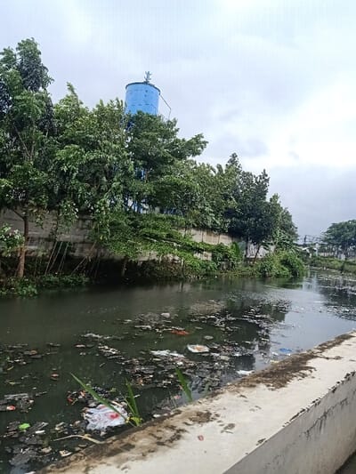 A murky canal overflowing with trash contrasts with the cloudy sky. A concrete retaining wall lines the canal, supporting a dense row of trees and a blue water tower perched on the rise behind them. The water is heavily polluted with scattered debris. The scene evokes a sense of environmental neglect and urban decay.
