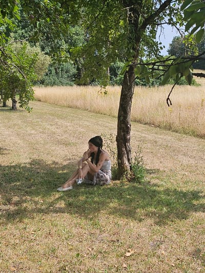 A young woman with dark hair, wearing a light-colored tank top and a patterned skirt, sits in the shade of a tree on a dry, grassy field. She is holding a white object in her hand and appears to be eating or examining it. The background consists of a large field of dry, golden grass and a line of trees. The image is taken outdoors on a sunny day. The location is Échebrune, France.
