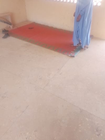 This image captures an interior room in Kanwa, Nigeria. The floor consists of light-toned, speckled terrazzo or concrete tiles with visible grout lines. A light-beige wall forms the background, featuring a narrow, horizontally slatted window positioned high on the wall. A rectangular red mat, bordered with a green and black pattern, is laid flat on the floor. The lower body of an individual, dressed in a flowing, light blue garment with a patterned hem, is visible. The person stands barefoot on the mat, facing away from the camera. A dark-colored object, consistent with a mobile phone, rests on the mat to the left of the individual. A pair of dark athletic shoes is placed on the tiled floor adjacent to the mat's left edge. In the far upper-left corner, a light-colored, possibly wooden, wall-mounted shelf is partially visible.