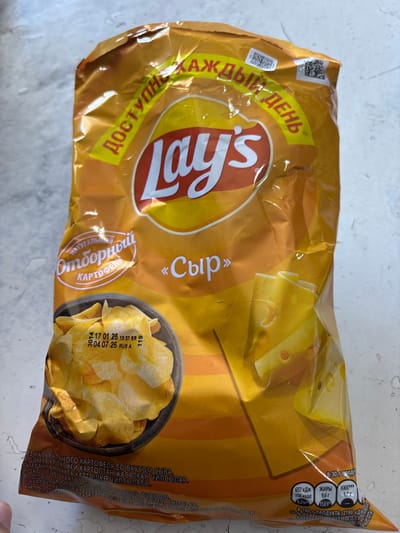 The scene depicts a single Lay's brand potato chip bag, predominantly orange and yellow, resting on a light gray, textured surface resembling a countertop. The bag is crinkled in several areas, indicating prior handling. Prominent Cyrillic text on the bag identifies the flavor as "Сыр" (Cheese) and features the slogan "ДОСТУПНЫЙ КАЖДЫЙ ДЕНЬ" (Available Every Day). Graphics include a dark bowl filled with potato chips on the lower left and several overlapping slices of cheese on the lower right. A printed date stamp on the image of the chips indicates "17 01 26 13:37 SR" (likely production date and time: January 17, 2026, 13:37) and "04 07 26 RUS A" (likely best-before date: July 4, 2026, manufactured in Russia, Batch A). Nutritional information is visible on the bottom right corner. No individuals or specific actions are discernible. The lighting is even, suggesting an indoor environment. This image is documented in the city of Lehvaz, Armenia.
