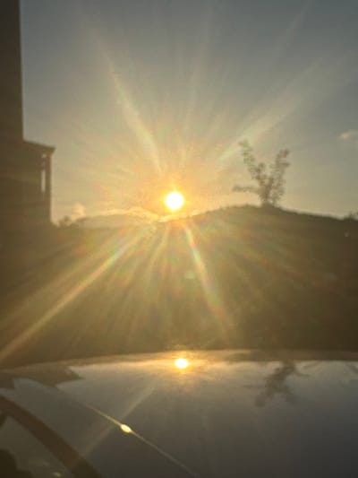 The image captures a sunset scene in Ammouliani, Greece. The sun, a bright orb, is positioned slightly left of the center and is emitting strong rays that create a halo effect. In the foreground, a reflective surface, likely the hood of a car, mirrors the sunset and the surrounding environment. To the left, a dark, vertical structure, possibly a building or a part of a building, is visible. Behind the sun, a silhouetted landscape features a hill with a sparse, leafless tree on its crest. The sky is a gradient of light blue and yellow hues, with wispy clouds present. The overall image is characterized by the intense glare of the sun and its scattered light, giving it a warm and bright atmosphere.