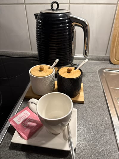 The image depicts a kitchen counter in Velbert, Germany, featuring items for preparing a hot beverage. A black ribbed electric kettle with a chrome handle is prominently positioned. To its left are two small ceramic containers with wooden lids and small spoons; one container is white and textured, the other is black and textured. A small wooden board supports these containers. In the foreground, a white teacup sits on a square saucer, with a single tea sachet, pink and labeled "WESTMINSTER STARTE DURCH", placed on the saucer. A small spoon rests on the saucer next to the teacup. The kitchen counter is grey and speckled, with a black induction stovetop visible to the left and a stainless steel sink to the right. The background shows a tiled wall. There are no people visible in the image.