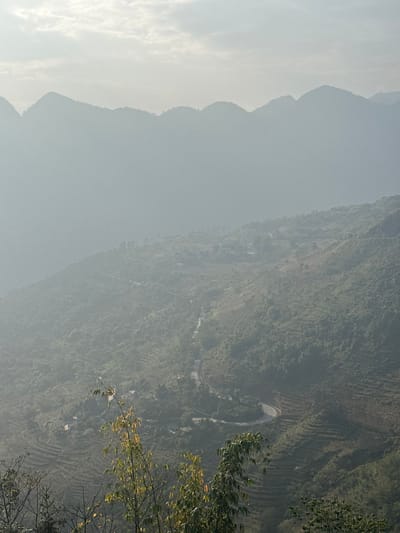This image depicts an expansive, hazy mountainous landscape within Minh Tân Commune, Vietnam. The background features multiple layers of rounded mountain peaks, appearing as blue-grey silhouettes against an overcast or diffusely lit pale sky. The mid-ground showcases a deep valley with a prominent, dark-colored, winding road traversing the slopes, visible with several turns and switchbacks. The hillsides are densely covered in green vegetation, interspersed with numerous agricultural terraces carved into the terrain, particularly on the lower right and mid-left slopes. Several small, light-colored structures, likely dwellings, are sparsely distributed across the mountainsides. The immediate foreground contains out-of-focus green and yellow-leafed foliage, including bamboo-like plants, partially obstructing the lower view. No individuals, animals, or vehicles are discernible in the scene. The overall illumination is soft and diffused due to atmospheric haze.