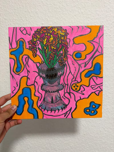 A square canvas features a vibrant, abstract painting of flowers in a vase. The background is a swirling mix of bright pink, orange, and blue shapes, outlined in thick black lines. Some of the blue shapes are irregular and amoeba-like, while others appear more structured, like two intertwined figures in the upper right corner. The flowers themselves are rendered in a variety of warm colors, including yellow, orange, and deep red, with green stems. They are clustered together in what appears to be a dark, textured vase with hints of purple and gray. The vase has a ribbed detail around its opening and a somewhat irregular shape. The overall style is bold and graphic, with a psychedelic feel. A hand is visible in the lower right corner, holding the canvas. The background wall is textured and white. No text is visible. The image appears to be taken indoors, with natural light illuminating the painting.