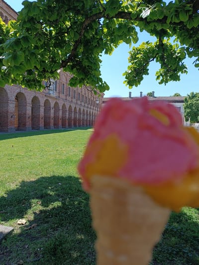 The image shows a blurred ice cream cone in the foreground. Behind it, there is a lawn, a long brick building with arched openings, and a tree providing shade. The image was taken in Mezzana Sant'Antonio, Italy.