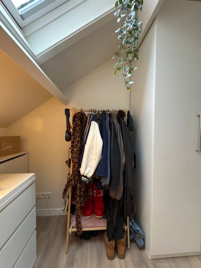 A leaning wooden rack holds clothes and shoes under a white cabinet. Garments include leather jackets, a blue jacket, a white and blue jacket, and a leopard-print coat. Footwear consists of brown boots, red boots, and blue sneakers. A black umbrella and silver necklace sit on the bottom shelf. A faux plant rests near a window. A partial view of a box with red lettering is visible beside a white, slatted structure.