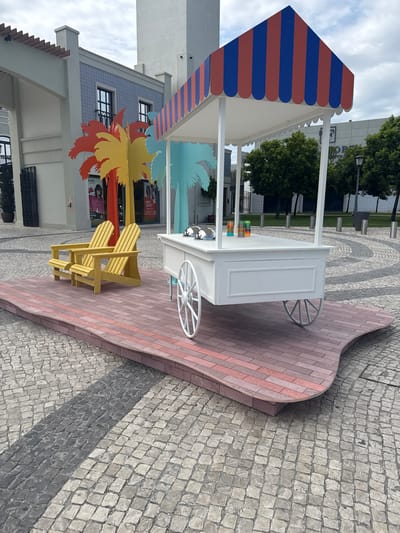 The image shows an outdoor scene, possibly a public plaza or walkway, with a decorative ice cream cart as the central object. The cart is white with a colorful striped awning in red and blue. It has decorative wheels, suggesting it's more for aesthetic purposes than practical mobility. In front of the cart are two bright yellow Adirondack chairs, inviting people to sit and relax. Behind the cart, a colorful cutout of palm trees in shades of yellow, orange, and red adds a tropical flair. The ground is paved with cobblestones, with a patterned inlay of alternating grey and light-colored stones. In the background, a modern building with large windows and some blue tiled sections is visible, along with trees and a cloudy sky, indicating it's daytime with overcast weather. The overall impression is a cheerful and inviting spot, likely designed for leisure and enjoyment, perhaps in a shopping area or a resort. There is no activity or people visible in the image, focusing solely on the setup and its environment. The location context provided is Alcochete, Portugal.