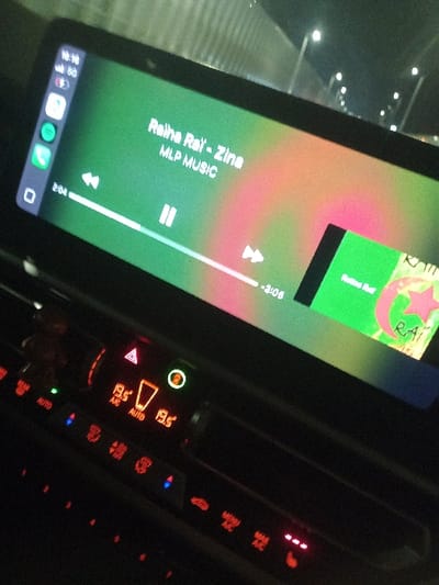 A car's infotainment system screen displays the music player interface. The song "Raina Rai - Zina" by MLP MUSIC is playing, with a green and pink album art. The playback progress is visible, showing 2:04 elapsed time and 3:05 remaining. To the right, the car's climate control panel glows with orange and red displays. Temperature settings are set to 19.5°C in auto mode, with various illuminated buttons controlling air circulation and heated seats.