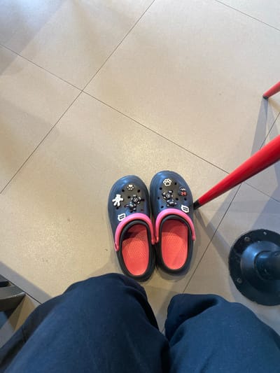 A pair of dark gray Crocs with pink trim and insoles sits on a tiled floor. The Crocs are adorned with several decorative charms, including a white paw print, a white teddy bear, and what appears to be a Mickey Mouse head. The scene is viewed from a high angle, looking down. To the left of the shoes, a red chair leg is visible, and to the right, a dark blue fabric, likely clothing, obscures part of the image. In the background, a black, circular base of what seems to be a table or stand is also present. The lighting suggests an indoor setting, possibly during the daytime. The overall impression is casual, with the focus on the footwear. No visible text is discernible on the Crocs or surrounding objects. The location context provided is Lhoksukon, Indonesia.
