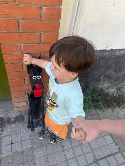 A young boy wearing a light blue t-shirt with a yellow and white truck graphic and orange shorts, with dark hair and fair skin, stands next to a brick wall. He is holding the hand of an adult. The boy is interacting with a black metal garden sculpture that resembles a dog, with red and white accents. The sculpture is mounted on a metal stand. The scene appears to be outdoors on a paved walkway, with some sparse greenery and a white and gray building in the background. The location is stated to be Temryuk, Russia.