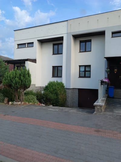 This is a medium-angle, eye-level, outdoor, full shot of a two-story white house with a dark brown garage door on the ground floor, located on a paved driveway in the city of Horažďovice, Czechia. The house has several dark brown framed windows on both floors. The ground floor of the house is partially clad in gray stone. A manicured green shrub is planted in front of the house, to the left of the entrance. The driveway is paved with interlocking concrete blocks, with a red patterned stripe running through it. In the background, to the right of the house, there is a blue trash bin, a stone retaining wall, and a dark brown front door with some hanging decorations. The sky above is blue with scattered white clouds. The scene appears to be a residential property.