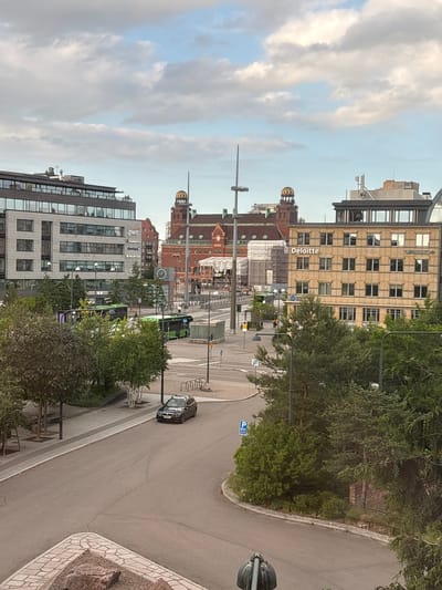 The image shows a street scene in Malmö, Sweden, likely near the central business district. In the foreground, a dark-colored sedan is parked on a paved road. The road curves to the right, leading towards a sidewalk lined with trees and landscaping.
In the midground, two green buses are visible on a street. One bus is stopped, and the other is in motion. A pedestrian is walking on the sidewalk to the right of the buses. Several modern office buildings are prominent in the background. One building on the left features the "PwC" logo. Another building on the right has the "Deloitte" logo. Across the street from the Deloitte building, a red brick building with a distinctive dome is visible. The sky above is partially cloudy, with patches of blue. The overall scene depicts a typical urban environment with traffic, pedestrians, and commercial architecture.
