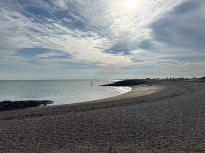 The image shows a wide, shingle beach leading to the sea, with a grey, slightly choppy sea under a bright, cloudy sky. In the distance, there are rock breakwaters and posts in the water, indicating a coastal defense or navigation aid. The beach itself is covered in dark grey pebbles and some patches of seaweed. The sky is dominated by a mix of white and grey clouds, with the sun shining through, creating bright, hazy areas. There are a few distant wind turbines visible on the horizon, suggesting an open, coastal landscape. The overall impression is a slightly overcast but bright day at a seaside location, likely St Osyth, United Kingdom, given the presence of coastal features and open horizons typical of the area. There are no people clearly visible in the foreground or mid-ground, although a few figures might be discernible in the distance on the beach.