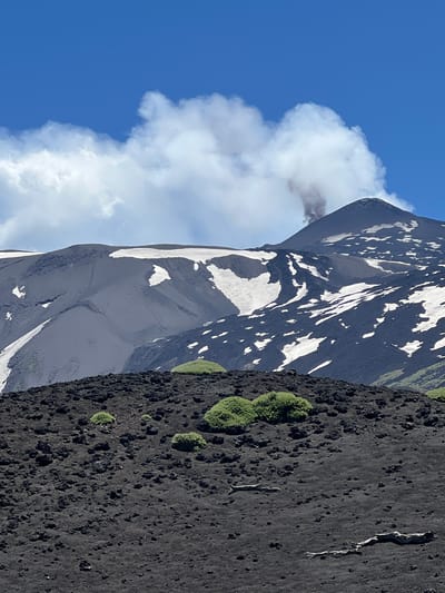 An outdoor, eye-level, medium shot captures the side view of Mount Etna, a volcano in Linguaglossa, Italy. The volcano's summit is visible on the right side of the frame, with a plume of smoke rising from its peak. The mountain's slopes are covered in dark volcanic rock and patches of snow, suggesting a high altitude. In the foreground, a rocky, dark grey landscape is dotted with small patches of green vegetation. The sky above is a clear, vibrant blue with fluffy white clouds scattered across it. The image is well-lit, indicating daylight. No people or animals are present in the scene.