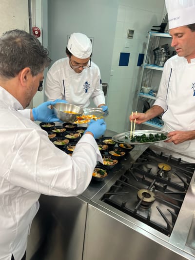 In a well-lit commercial kitchen located in Pozuelo de Alarcón, Spain, three male chefs are engaged in the assembly-line plating of individual food portions. The kitchen features white tiled walls with blue accent tiles, stainless steel countertops, and commercial kitchen equipment.
From left to right:
1. **Foreground Chef:** A male with medium-length dark grey hair, wearing a white chef's jacket and light blue disposable gloves. He holds a silver metal bowl containing an orange-yellow liquid, tilting it to pour into multiple small, black, individual serving ramekins arranged on the stainless steel counter. These ramekins already contain various ingredients, including noodles, mushrooms, and a distinct yellow circular component.
2. **Middle Chef:** A younger male wearing a white chef's jacket with blue trim, a white chef's hat featuring a blue and white "Le Cordon Bleu" emblem, and glasses. He holds a silver oval tray containing numerous segmented orange pieces, presenting them as part of the plating process.
3. **Right Chef:** A male with dark hair, wearing a white chef's jacket with blue trim and a tall white chef's hat bearing the "LE CORDON BLEU" text. He holds a silver oval tray filled with dark green leafy vegetables (likely spinach) in his left hand. His right hand uses wooden chopsticks to precisely transfer these vegetables into the individual black ramekins on the counter.
In the background, a blue-shelved metal rack holds various kitchen utensils and metal bowls. A red fire alarm pull station is visible on the left wall. A four-burner gas range, not in operation, is integrated into the stainless steel counter on the right. The overall scene depicts a professional and organized culinary operation.