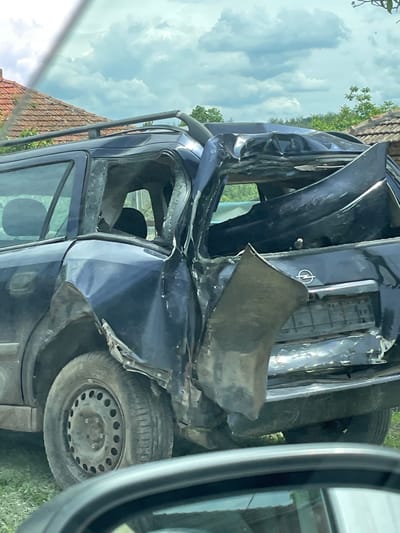 The image depicts a heavily damaged blue Opel station wagon, likely in Osenets, Bulgaria, based on the surrounding context. The vehicle appears to have sustained significant structural damage, particularly to the driver's side and front. The driver-side door is detached and crushed. The front is severely compromised with crumpled metal. The windows are broken. The car rests on its wheels, with one tire visible. The environment consists of a building with a red tile roof and foliage. The image was captured from inside another vehicle, with a portion of the side mirror visible in the frame. There are no people visible in the immediate vicinity of the damaged car.