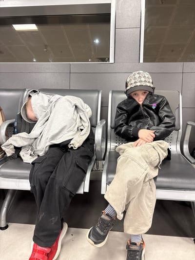 Two children are sitting on gray airport-style seats in what appears to be an airport waiting area. The child on the left is asleep, with their head covered by a gray hoodie and draped over their lap. They are wearing black cargo pants and red sneakers. The child on the right is awake and looking towards the camera. They are wearing a black leather jacket, a patterned baseball cap, beige pants, striped socks, and black sneakers. The background shows a modern, gray tiled wall and a reflective ceiling with lighting fixtures. The floor is a light gray material.