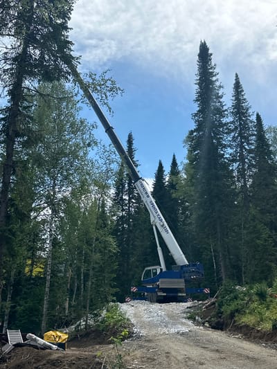 A large blue and white crane is positioned on a dirt road in a dense forest. The crane's boom extends upwards, reaching towards the top of a tall evergreen tree. The tree appears to be leaning or in the process of being lowered. The area around the crane is uneven, with piles of gravel and dirt. Various equipment, including what appears to be lumber and a yellow bag, are scattered on the ground near the base of the slope. The background is filled with a canopy of tall evergreen trees. The sky is partly cloudy. The scene is indicative of tree removal or logging operations. The location is identified as Sheregesh, Russia.