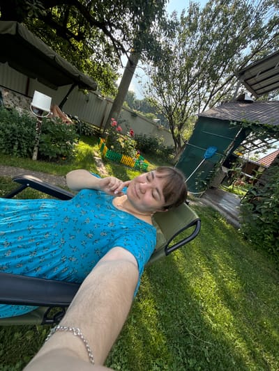 A young woman sits in a green outdoor chair, taking a selfie. She is wearing a blue floral dress and has a thoughtful expression, with her eyes closed and a finger gently touching her cheek. The scene is a backyard in Nizhne-Chufichevo, Russia, on a sunny day. Lush green grass surrounds the chair, with sunlight dappling through the leaves of a large tree. In the background, there's a wooden fence, a small building with a green tarp, and a garden bed with roses. A blue butterfly net leans against the building. The overall atmosphere is relaxed and peaceful, suggesting a leisurely afternoon outdoors.