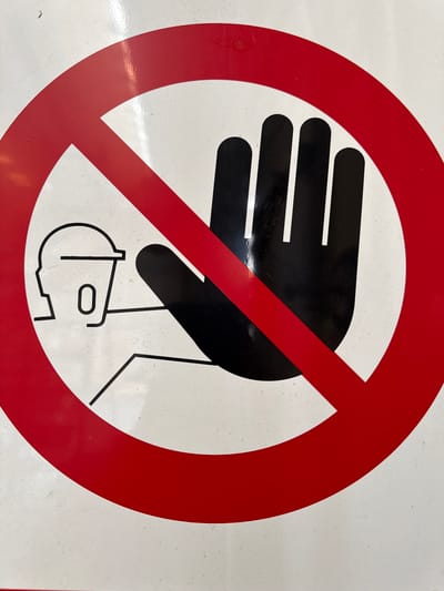 A red prohibition circle with a diagonal slash intersects a black silhouette of a hand raised in a stop gesture. To the left, a simplified black outline of a person's head, wearing what appears to be a hard hat, is positioned behind the hand. The overall graphic conveys a "do not touch" or "no entry" message, likely a safety or access restriction. This sign is affixed to a white surface, which shows signs of minor wear and scuffing. The image was captured in Fontevivo, Italy.